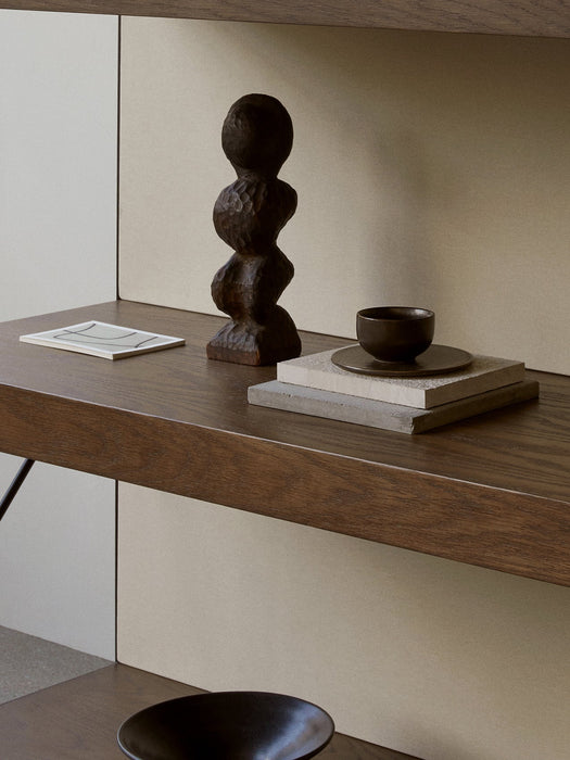 A close-up of a dark wood shelf styled with a carved wood sculpture, a small ceramic bowl on stone coasters, and an art card.