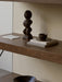 A close-up of a dark wood shelf styled with a carved wood sculpture, a small ceramic bowl on stone coasters, and an art card.