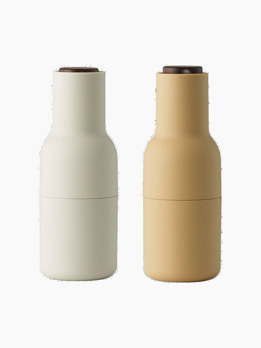 A pair of Audo Bottle Grinders, one barley and one off-white, both with dark walnut wood lids. Shot on a black background.