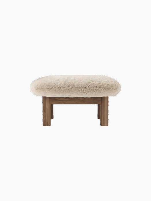 Back view of the Brasilia Ottoman, featuring a plush, cream sheepskin cushion on a wooden base with four cylindrical legs.