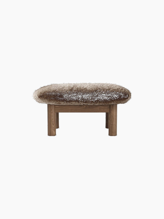 A front view of the Brasilia Ottoman, featuring a plush, brown sheepskin top and four robust, light-toned wooden legs.