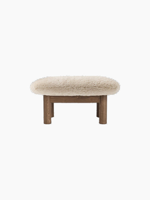 Front view of the Brasilia Ottoman, Sheepskin, featuring a plush, off-white cushion and four robust, cylindrical wooden legs.
