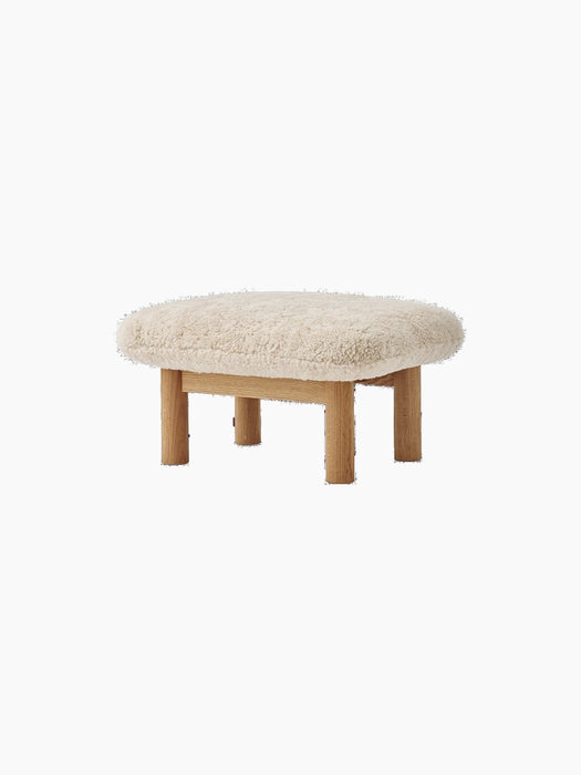 Angled view of the Brasilia Ottoman with a plush, off-white sheepskin cushion and four cylindrical legs in natural oak.