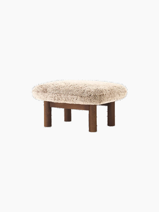 Brasilia Ottoman with a plush Sheepskin Root cushion and four robust, cylindrical legs in dark stained oak, at an angle.