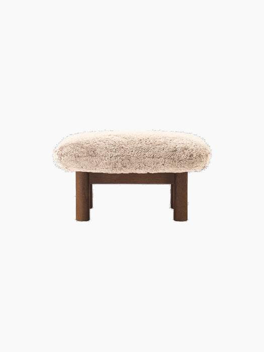 The Brasilia Ottoman with a plush Sheepskin Root cushion on a dark-stained oak frame and four robust legs.
