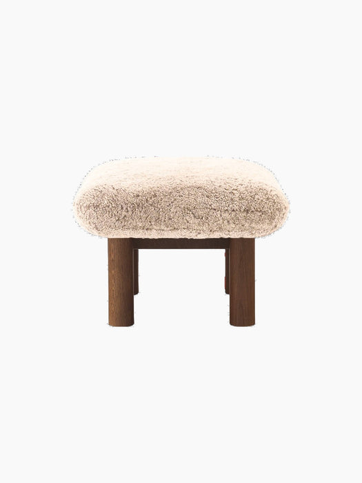 A side profile of the Brasilia Ottoman, showcasing its plush, light brown sheepskin seat atop a robust dark stained oak base.