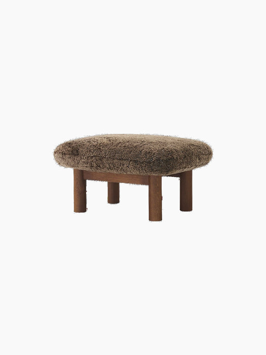 The Brasilia Ottoman with a fluffy brown sheepskin cushion on a dark stained oak base with four thick, cylindrical legs.