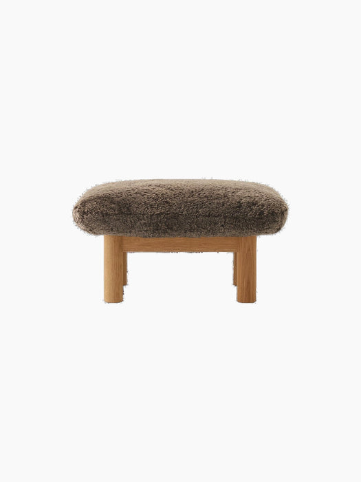 A front view of the Brasilia Ottoman, featuring a plush, brown sheepskin cushion atop a natural oak frame with robust legs.