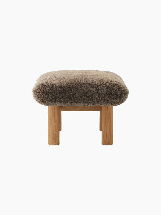 Side profile of the Brasilia Ottoman, featuring a thick, brown sheepskin cushion and robust cylindrical legs in natural oak.