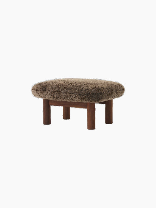 The Brasilia Ottoman in walnut wood with a brown sheepskin upholstery, seen from an angle against a black background.