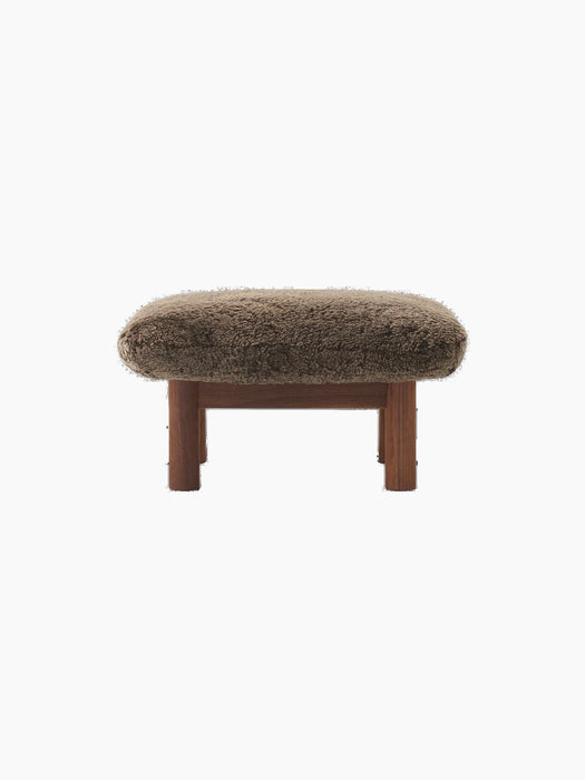A front-on view of the Brasilia Ottoman, upholstered in brown sheepskin, with a dark walnut frame and four thick legs.