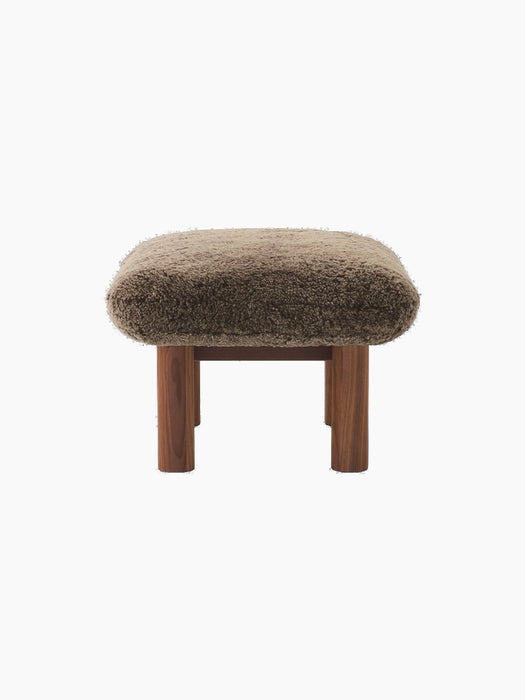 Side view of the Brasilia Ottoman, featuring a plush brown sheepskin cushion and robust, cylindrical legs in walnut wood.