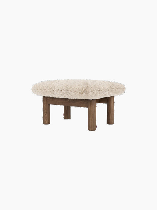 Angled view of the Brasilia Ottoman with a plush sheepskin cushion and four robust, cylindrical wooden legs.