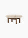 Angled view of the Brasilia Ottoman with a plush sheepskin cushion and four robust, cylindrical wooden legs.