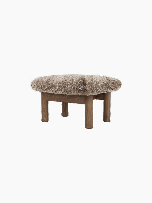 Brasilia Ottoman with a plush sheepskin cushion in a light brown hue, supported by four robust, cylindrical wooden legs.