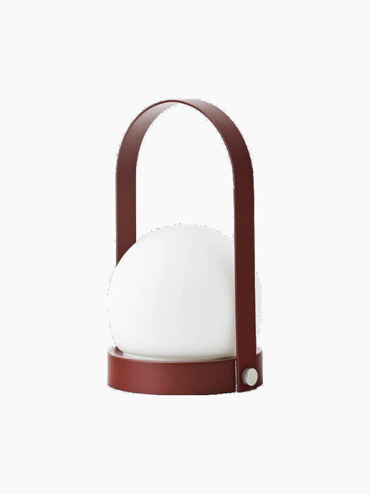 The Carrie Table Lamp in Burned Red, featuring a basket-like base, a matching arched handle, and a central white glass orb.