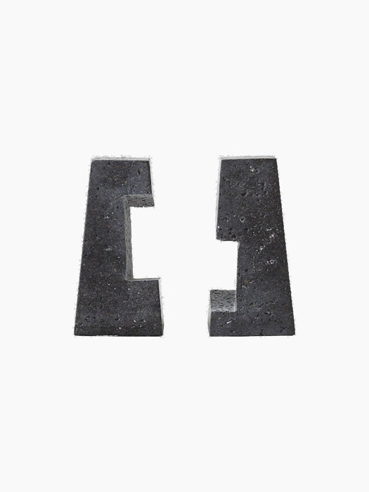 A pair of sculptural Converge Book Ends made from dark, porous lava stone, standing apart against a solid black background.