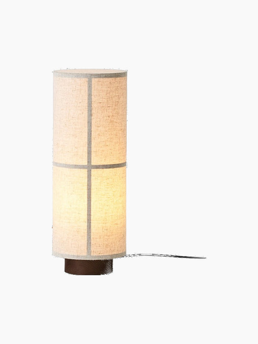 The Hashira Table Lamp in Raw, seen from an angle. The cylindrical linen shade is lit, sitting atop a dark wood base.