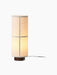 The Hashira Table Lamp in Raw, seen from an angle. The cylindrical linen shade is lit, sitting atop a dark wood base.