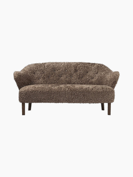 A front view of the Ingeborg Sofa in dark brown Sahara sheepskin, featuring a button-tufted back and dark stained oak legs.