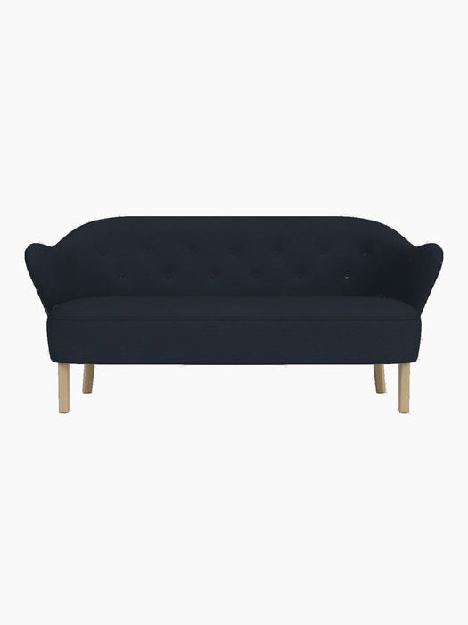 A front-on view of the Ingeborg Sofa. It features dark blue button-tufted upholstery, curved armrests, and natural oak legs.