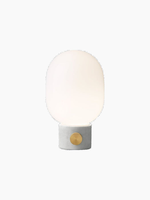 Front view of the JWDA Table Lamp, Small, with a light grey concrete base, brass dimmer knob, and lit opal glass shade.