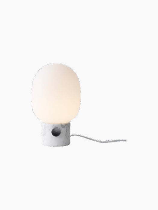 A lit JWDA Table Lamp, Large with a white Carrara marble base, black dimmer switch, and glowing opal glass shade.