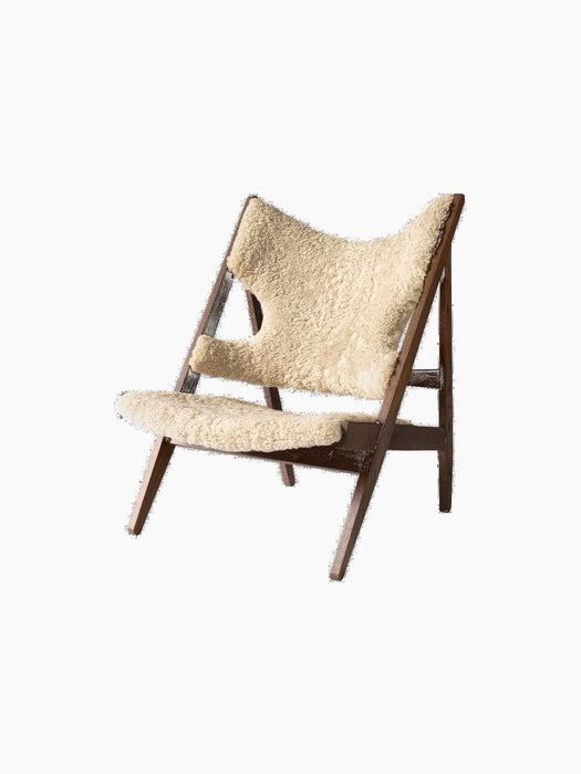 The Knitting Lounge Chair shown at an angle, with a dark stained oak frame and natural sheepskin upholstery