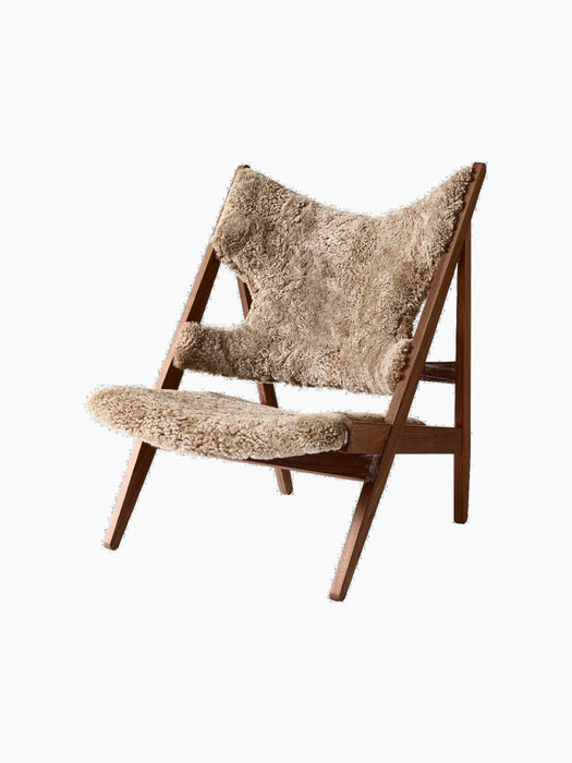 The Knitting Lounge Chair has a dark walnut A-frame with a seat and winged backrest upholstered in textured beige sheepskin.