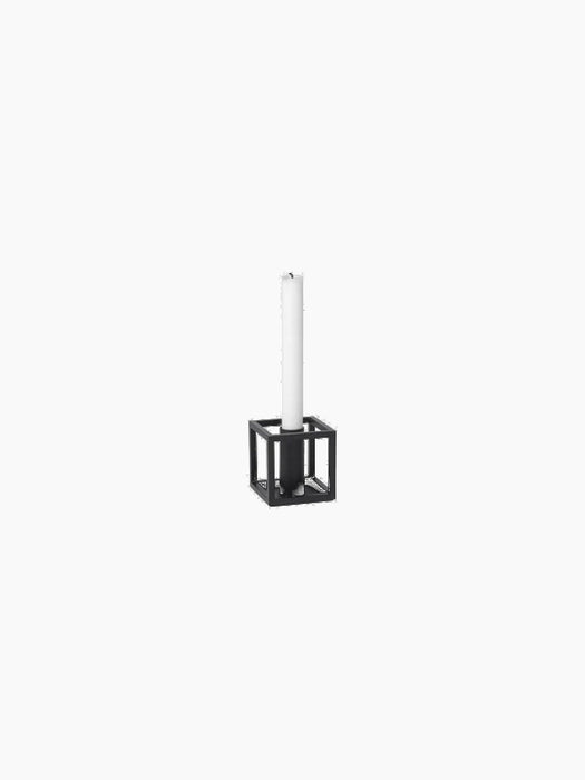 The Kubus 1 candle holder in black, a cube-shaped metal frame holding a single tall white candle against a black background.