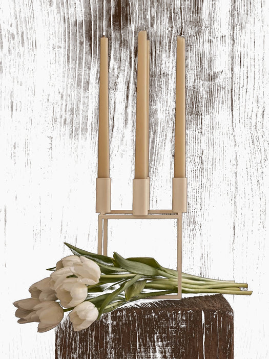 A white Kubus 4 candle holder with three cream candles on a dark wood surface with white tulips against a dark wood wall.