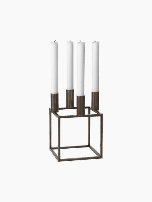 A Kubus 4 candle holder in burnished copper finish, featuring a geometric cube-shaped frame holding four tall white candles.