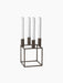 A Kubus 4 candle holder in burnished copper finish, featuring a geometric cube-shaped frame holding four tall white candles.