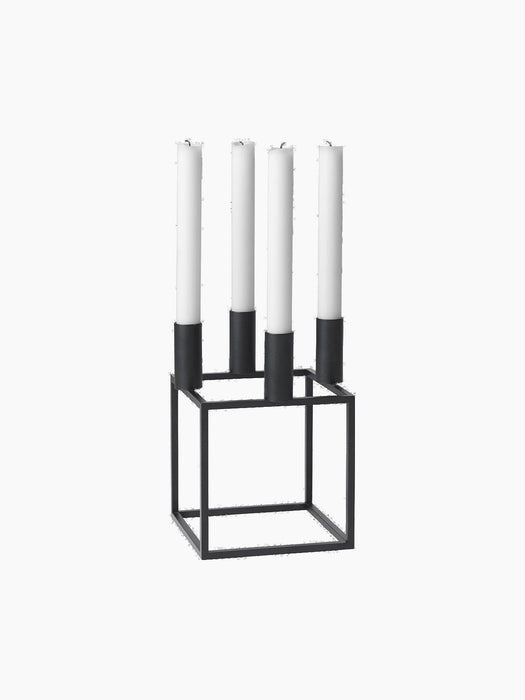 A black, geometric Kubus 4 candle holder with a square base, holding four tall white unlit candles, on a black background.