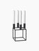 A black, geometric Kubus 4 candle holder with a square base, holding four tall white unlit candles, on a black background.