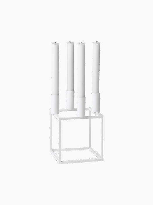 A white Kubus 4 candle holder with a geometric cube base, holding four tall, white, unlit candles against a black background.
