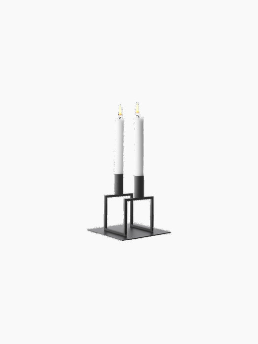 A minimalist, black metal candle holder with a square base and two geometric frames, holding two tall, lit white candles.