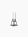 A minimalist, black metal candle holder with a square base and two geometric frames, holding two tall, lit white candles.