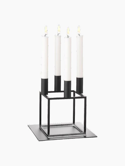 A black Kubus 4 candle holder with four lit white candles, sitting on a black Kubus Base, against a black background.