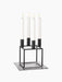 A black Kubus 4 candle holder with four lit white candles, sitting on a black Kubus Base, against a black background.