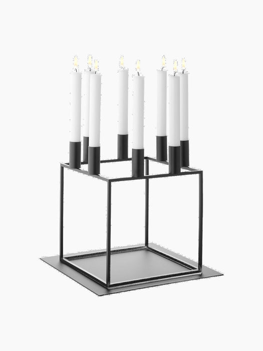 A black metal Kubus 8 candle holder with eight lit white candles, shown on the matching square Base for Kubus 8.