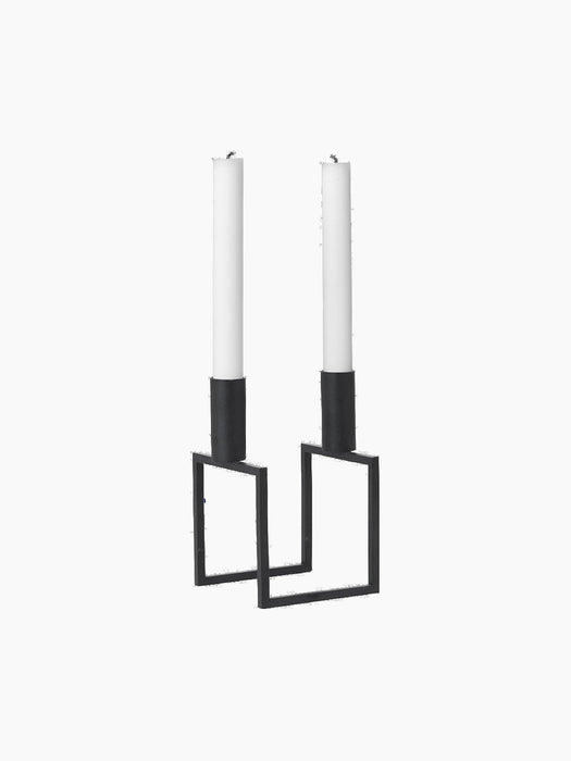 A black, geometric Kubus Line candle holder with two tall white candles, viewed from an angle against a black background.