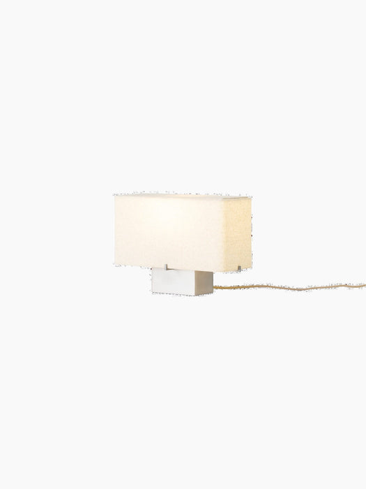 An angle view of the illuminated Nonna Table Lamp, featuring a rectangular sand-coloured textile shade on an aluminium base.
