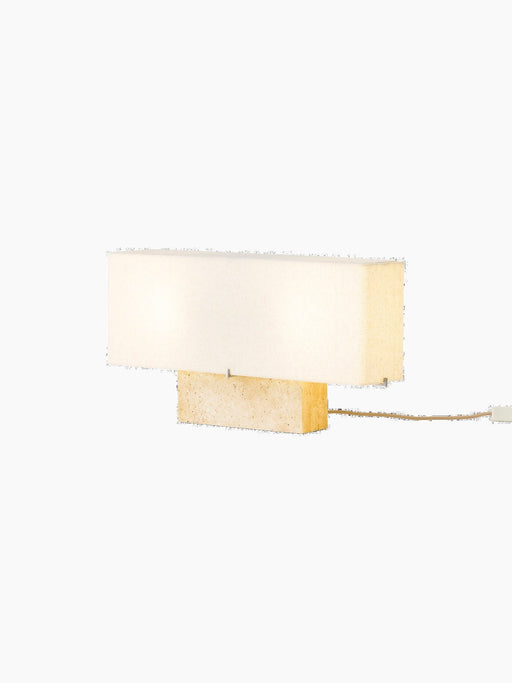 Angled view of the Nonna Table Lamp in Travertine, illuminated to show its rectangular stone base and long, fabric lampshade.