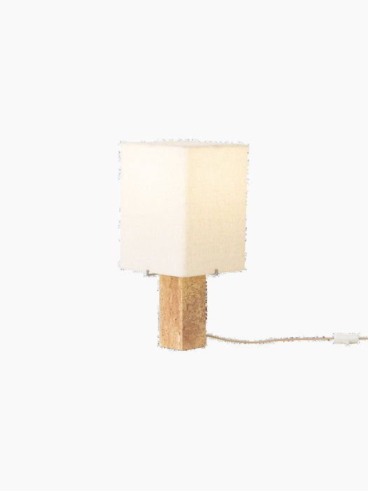 Angled view of the lit Nonna Table Lamp, featuring a square travertine base and a warm, glowing sand-coloured linen shade.