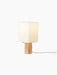 Angled view of the lit Nonna Table Lamp, featuring a square travertine base and a warm, glowing sand-coloured linen shade.