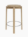A front view of the Passage Counter Stool in natural oak, showing its round seat, three legs, and stainless steel footrest.