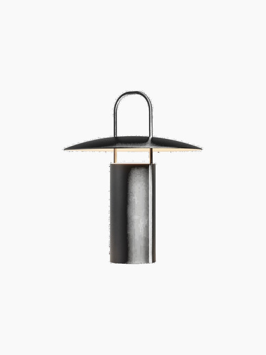 A black Ray Table Lamp, Portable, shown from an angle. It is turned on, casting a warm, soft glow from under its wide shade.