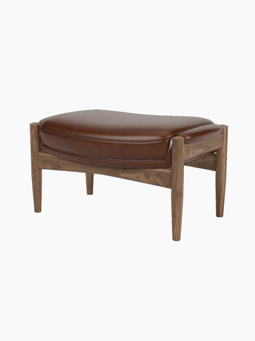 A three-quarter view of The Seal Ottoman, featuring a rich walnut wood frame and a plush, contoured brown leather cushion.