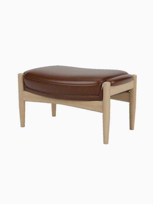 A side-profile view of The Seal Ottoman, showing its plush brown leather cushion and natural oak frame with tapered legs.